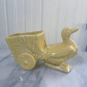 May include: A light yellow ceramic planter shaped like a duck pulling a cart. The cart has a rectangular opening for plants and a wheel on one side. The duck has a long beak and is standing on two feet.