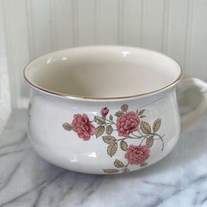 May include: Cream-colored ceramic chamber pot with a gold rim and handle. The pot is decorated with pink rose floral patterns and green leaves. The interior is a lighter cream color.
