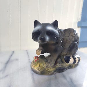 May include: A ceramic raccoon figurine with a black and white face and a striped tail. The raccoon is posed on a bed of leaves with red berries. The figurine is painted in shades of gray, brown, and white.