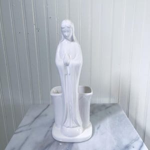 May include: A white ceramic statue of a woman in a flowing robe, holding a closed bud. The statue is part of a vase with two openings. The figurine is on a marble surface.