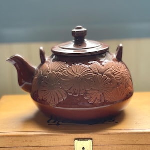 May include: A brown ceramic teapot with a lid and a spout. The teapot features a floral design embossed on its surface. The teapot is sitting on a wooden surface. The teapot has a handle on each side.