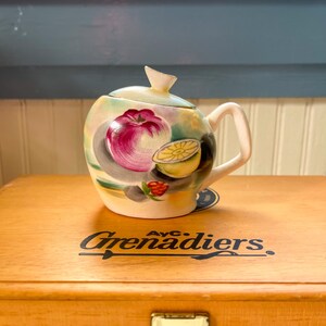 May include: A white ceramic teapot with a lid, painted with a colorful fruit design. The teapot has a handle and a spout. The teapot is sitting on a wooden box with the text "AYC Grenadiers" printed on it.