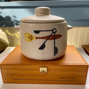 May include: A white ceramic casserole dish with a lid. The dish has a brown, yellow, and black design of a fish with a fork and spoon. The dish is sitting on a wooden box.