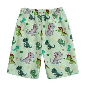 Puede incluir: Shorts verdes con un patrón de dinosaurios de dibujos animados en varios colores. Los dinosaurios están sobre un fondo verde claro.