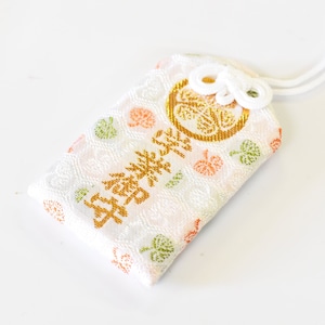 Puede incluir: Amuleto de tela blanca con estampado floral dorado y rosa y letras doradas que dicen "幸福祈願". El amuleto está atado a una cuerda blanca.