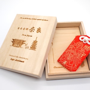 May include: A red and gold Japanese Omamori charm in a wooden box. The charm is embroidered with a figure and the text "勝" which means "victory". The box is engraved with the text "日本台都奈良 Nara Japan" and "Omamori lucky charm for you Saiga Hashimode".