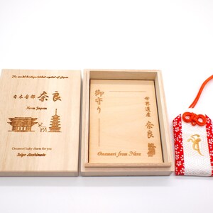 Op de afbeelding: Een houten doos met een rode en witte stoffen charme erin. De doos is gegraveerd met Japanse karakters en de woorden "Nara Japan" en "Omamori from Nara".