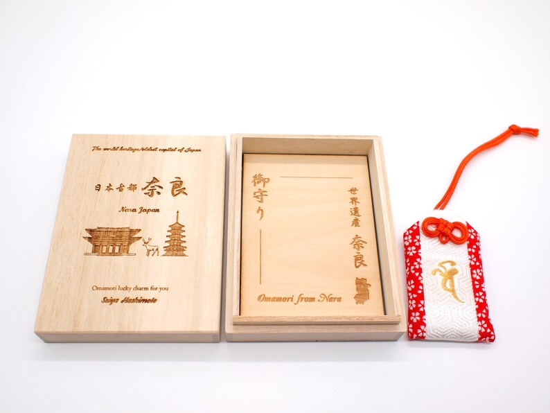Op de afbeelding: Een houten doos met een rode en witte stoffen charme erin. De doos is gegraveerd met Japanse karakters en de woorden "Nara Japan" en "Omamori from Nara".