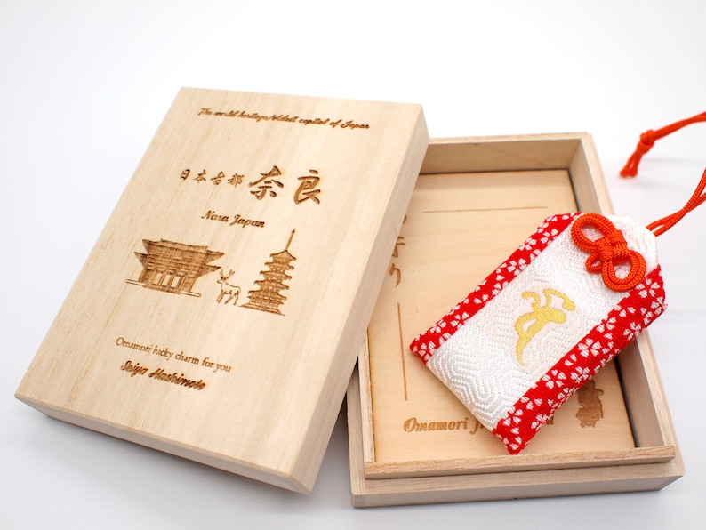 Op de afbeelding: Een houten doos met een rode en witte stoffen charme erin. De doos is gegraveerd met de woorden "Nara Japan" en een afbeelding van een tempel. De charme is een traditionele Japanse "Omamori"-geluksbrenger.