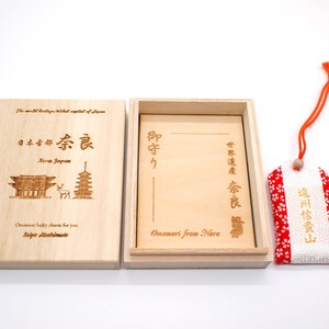 Op de afbeelding: Een houten doos met een rood-witte stoffen charme erin. De doos is gegraveerd met Japanse karakters en de woorden "Nara Japan" en "Omamori from Nara". De charme is een traditionele Japanse geluksbrenger.