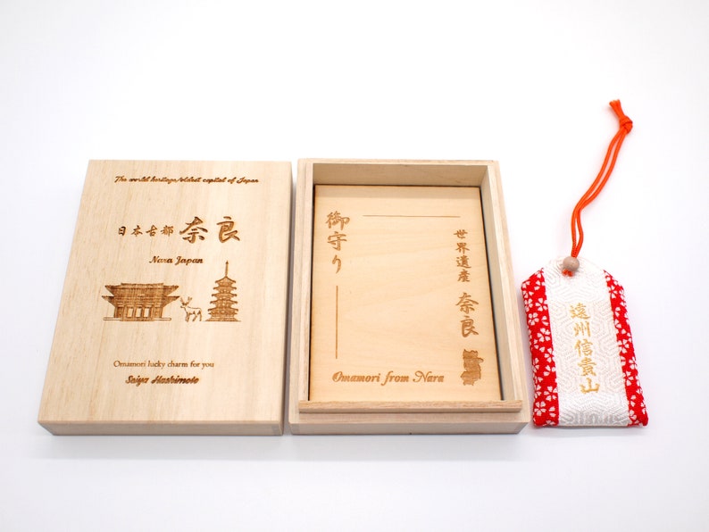 Op de afbeelding: Een houten doos met een rood-witte stoffen charme erin. De doos is gegraveerd met Japanse karakters en de woorden "Nara Japan" en "Omamori from Nara". De charme is een traditionele Japanse geluksbrenger.