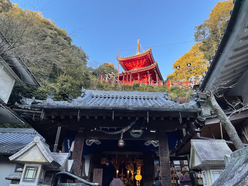 Op de afbeelding: Een rode pagode-stijl tempel met een zwart betegeld dak, gezien vanaf beneden. De tempel ligt op een heuvel en is omgeven door bomen. De tempel is een populaire toeristische bestemming in Japan.