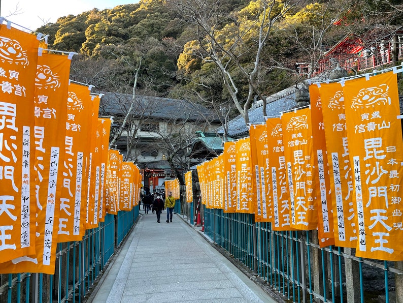 Op de afbeelding: Een lange gang met aan weerszijden oranje vlaggen met Japanse tekst en een tijgersymbool. De vlaggen hangen aan een blauw metalen hek. De gang leidt naar de ingang van een tempel.