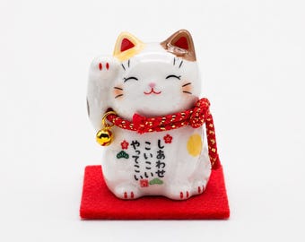May include: A white ceramic Maneki Neko figurine with a raised paw, adorned with a red collar and a gold bell. The cat has brown and yellow accents and sits on a red square. Japanese text is visible on the cat's body.