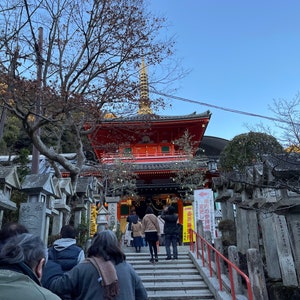 Op de afbeelding: Een rode en gouden Japanse tempel met een pagodadaken en een stenen trap die naar de ingang leidt. Mensen lopen de trap naar de tempel op.