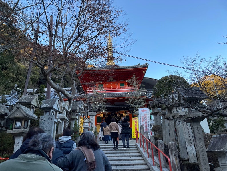 Op de afbeelding: Een rode en gouden Japanse tempel met een pagodadaken en een stenen trap die naar de ingang leidt. Mensen lopen de trap naar de tempel op.