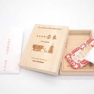 以下が含まれることがあります： 赤い布製の御守が入った小さな木製の箱。箱には「日本の世界遺産の首都奈良日本」と「あなたのための御守のお守り、とても縁起が良い」と書かれています。御守は金色の刺繍が施され、白い房が付いています。