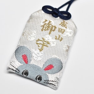 Pode incluir: Um amuleto omamori japonês com um design de rato. O amuleto retangular tem um fundo cinza claro e caracteres japoneses dourados. O rato tem orelhas cinzentas com detalhes vermelhos e olhos pretos. Um cordão azul marinho está preso.