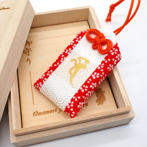 Op de afbeelding: Een witte en rode Japanse charme, genaamd "Omamori", in een houten doos. De charme heeft een gouden inscriptie en een rode strik. De doos is beschreven met Japanse karakters.