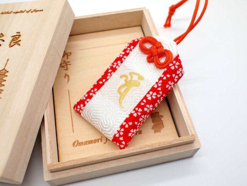 Op de afbeelding: Een witte en rode Japanse charme, genaamd "Omamori", in een houten doos. De charme heeft een gouden inscriptie en een rode strik. De doos is beschreven met Japanse karakters.