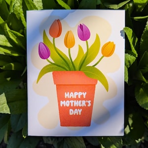 May include: A greeting card with a floral illustration. The card features a terracotta-colored flower pot with the text "HAPPY MOTHER'S DAY" in white. The pot holds tulips in shades of orange, yellow, and purple, with green leaves.