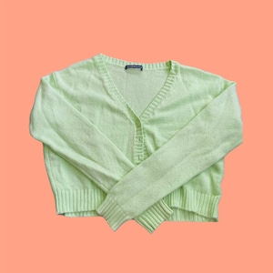 May include: A light green cropped cardigan with a V-neck and button closure. The cardigan has a ribbed texture and is made of a soft, lightweight material.