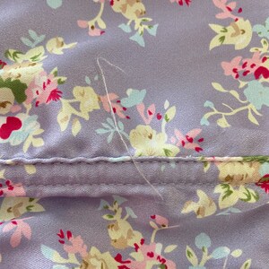 Pode incluir: Um close-up de um tecido roxo com padr&atilde;o floral. O tecido tem uma costura que atravessa o meio e um &uacute;nico fio branco pendurado na costura.