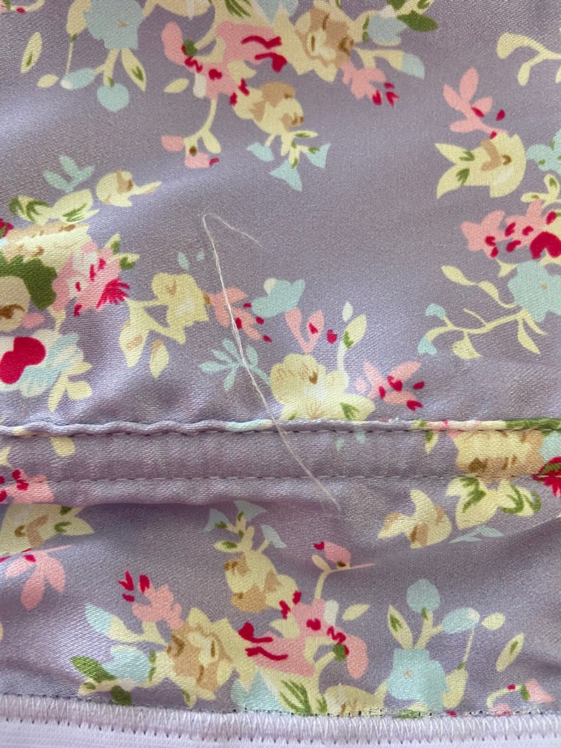 Pode incluir: Um close-up de um tecido roxo com padr&atilde;o floral. O tecido tem uma costura que atravessa o meio e um &uacute;nico fio branco pendurado na costura.