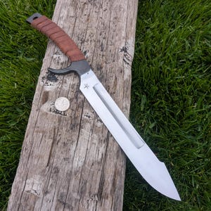 May include: A large, silver-colored knife with a brown leather-wrapped handle. The blade is wide and has a distinctive shape. A small coin is next to the knife on a weathered wooden surface, with green grass in the background.