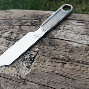 K&ouml;nnte beinhalten: Ein silbernes Messer mit schwarzem Griff und einem kleinen Baumlogo auf der Klinge. Das Messer liegt auf einem St&uuml;ck verwittertem Holz.