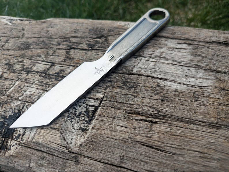 K&ouml;nnte beinhalten: Ein silbernes Messer mit schwarzem Griff und einem kleinen Baumlogo auf der Klinge. Das Messer liegt auf einem St&uuml;ck verwittertem Holz.