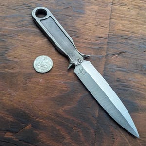 May include: A silver-colored dagger with a pointed blade and a handle with a hole at the end. The dagger has a cross symbol etched into the blade. A quarter is next to the dagger for scale. The dagger is on a wooden surface.