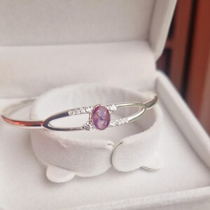 May include: A silver bangle bracelet with a purple oval gemstone and small clear stones set in the band.