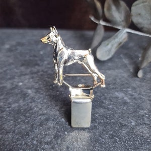以下が含まれることがあります： 銀色の金属製の犬の置物が、銀色の金属製のクリップに取り付けられています。犬は後ろ足で立ち、前足を上げています。
