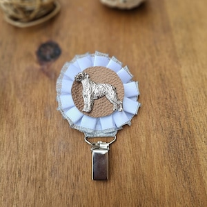May include: A silver dog silhouette on a brown leather circle, surrounded by a white and silver ruffled fabric rosette. The rosette is attached to a silver clip.