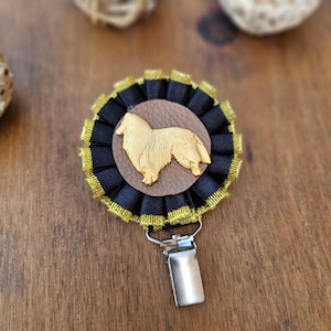 Op de afbeelding: Een gouden hondensilhouet op een bruine lederen achtergrond, omringd door een zwarte en gouden stoffen rozet met een gouden clip.