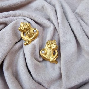 Può includere: Un paio di orecchini color oro, ciascuno con una testa di animale scolpita con un accento di perla. Gli orecchini sono appoggiati su uno sfondo di tessuto grigio chiaro.
