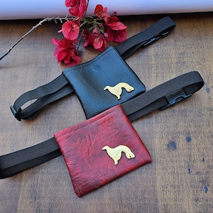 Puede incluir: Dos bolsas de cuero negro para golosinas de perro con correas ajustables y una silueta dorada de un perro en cada bolsa.