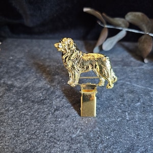 Może przedstawiać: Złota metalowa figurka psa przymocowana do złotego metalowego klipsa. Pies stoi na tylnych łapach i ma długi, puszysty ogon.