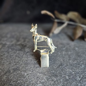 May include: A silver-coloured metal clip featuring a detailed dog figurine. The dog is standing, with its ears perked up. The clip is attached to the dog's underside, set against a dark background.