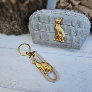 Può includere: Una pochette con zip in similpelle grigia con un design a forma di gatto dorato. Un portachiavi a forma di gatto dorato abbinato con un cinturino in pelle grigia.