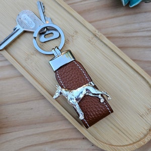 May include: A brown leather keyring with a silver dog charm. The keyring has a silver key ring and a silver loop.