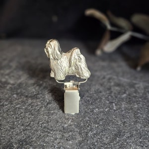 Pode incluir: Um clipe de metal prateado com uma figura detalhada de cão Shih Tzu. O cão é retratado com pelo longo e esvoaçante e fica em cima de um mecanismo de clipe retangular. O clipe foi projetado para segurar itens.