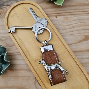Può includere: Un portachiavi in ​​cuoio marrone con un ciondolo a forma di cane argentato e un anello portachiavi argentato. Il portachiavi è su una superficie di legno.