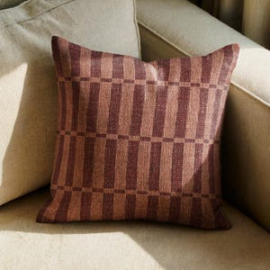 May include: A square throw pillow featuring a woven fabric texture with a repeating geometric pattern of vertical dark maroon and light copper-toned stripes, intersected by horizontal bands that create a structured, grid-like visual effect.