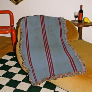 May include: A rectangular woven throw blanket featuring a dusty blue base color with two prominent vertical dark red stripes running down the center. The blanket has a textured appearance and is finished with a decorative fringe along all four edges.
