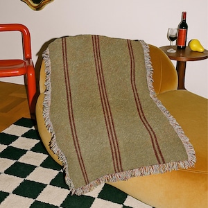 May include: A rectangular woven throw blanket featuring a textured olive green base with vertical dark red stripes and a multi-coloured fringed edge. The fabric has a visible knit construction with a soft, slightly fuzzy appearance throughout the surface.