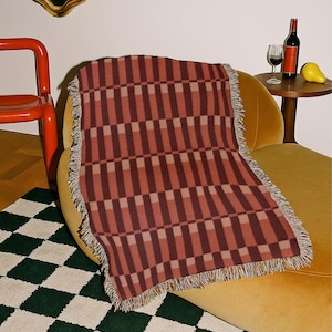 May include: A rectangular woven throw blanket featuring a repeating geometric pattern of vertical bars in shades of terracotta, dark maroon, and muted pink. The blanket has a textured surface and frayed edges along the sides and ends.