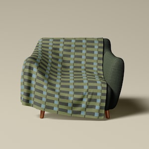 May include: A throw blanket featuring a repeating geometric pattern of vertical olive green and dark charcoal rectangles separated by thin light blue stripes, draped over a dark green armchair with wooden legs against a neutral background.