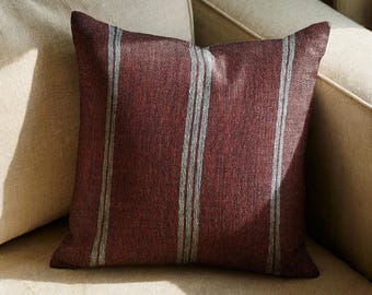Può includere: Un cuscino quadrato in tessuto rosso bordeaux intenso con una finitura testurizzata. Il cuscino è decorato con tre strisce verticali di colore grigio chiaro, ciascuna composta da due sottili linee parallele che corrono lungo il centro.